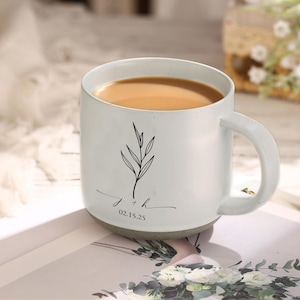 May include: White ceramic mug with a handle, filled with a light brown liquid. The mug features a black botanical illustration and the inscription "j + h 02.15.25". The mug has a gray base and is set on a light-colored surface.