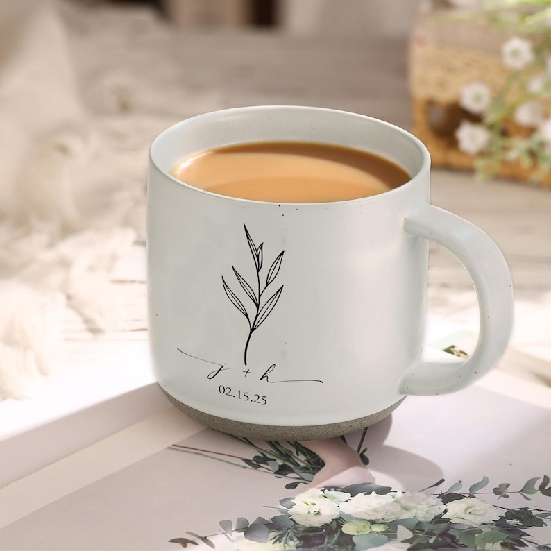 May include: White ceramic mug with a handle, filled with a light brown liquid. The mug features a black botanical illustration and the inscription "j + h 02.15.25". The mug has a gray base and is set on a light-colored surface.
