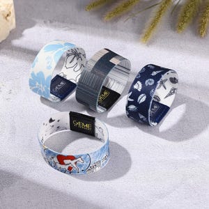 May include: Four fabric bracelets with different patterns. One has a blue floral design, another has a striped pattern, and a third features seashells. The fourth bracelet has a horse and cityscape design. Each bracelet has a black tag with the text "GEME AL JEWELRY".
