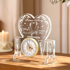 May include: A clear acrylic clock with a heart-shaped top, inscribed with "Forever & Always" and "Always Forever in my Heart." The clock face is white with gold trim and Roman numerals. The word "LOVE" is spelled out in clear acrylic letters.