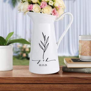 Puede incluir: Una jarra de cerámica blanca con asa, llena de un ramo de rosas blancas y rosas. La jarra tiene un diseño botánico negro y las iniciales "j + h" con la fecha "02.15.25" debajo.