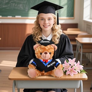 May include: A smiling graduate in a black gown holds a teddy bear dressed in a miniature graduation outfit. The bear has a small cap and gown with "Class of 2025" on it. Pink lilies are on the desk. The bear's cap says "Samme".