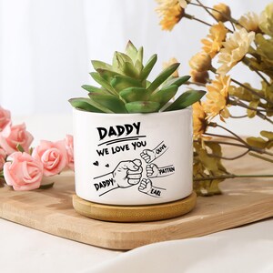 May include: A white ceramic planter with the text "DADDY WE LOVE YOU" and illustrations of fists. The planter holds a green succulent plant and sits on a wooden coaster. The names "OLIVE", "PATTEN", and "EARL" are also on the planter.