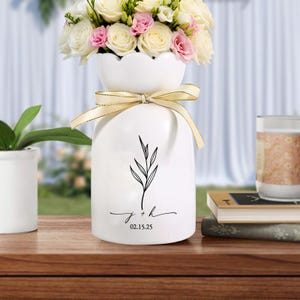 May include: White ceramic vase with a scalloped top, tied with a gold ribbon, and adorned with a black botanical illustration. The vase holds a bouquet of white and pink roses. The vase has the initials "j + h" and the date "02.15.25" printed on it.