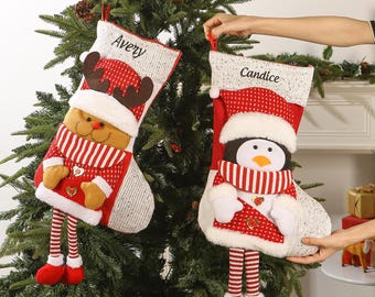 Op de afbeelding: Twee kerstsokken hangen aan een boom. Eén is een rendier met de naam "Avery" en de andere is een pinguïn met de naam "Candice". Beide sokken zijn rood, wit en beige met decoratieve details.