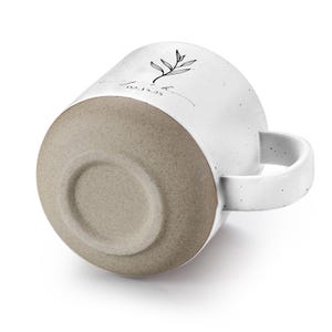 May include: White ceramic mug with a speckled finish, featuring a black botanical illustration and text. The bottom of the mug is a light brown color. The handle is white. The text on the mug reads "Love" and "02.15.23".
