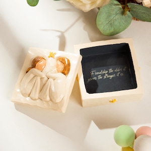 Puede incluir: Una caja de recuerdos de color crema con una tapa que presenta una escultura en relieve de dos figuras. La caja está abierta, revelando un interior oscuro con la inscripción "Friendship the older it grows the stronger it is."