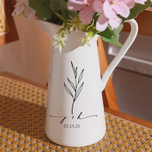 May include: White ceramic pitcher with a black leaf design and the initials "j + h" with the date "02.15.25". The pitcher is filled with pink and white flowers and green leaves. The pitcher has a handle and a spout.
