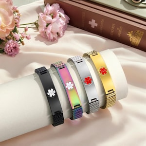 Puede incluir: Cinco pulseras de alerta médica en negro, iridiscente, plateado y dorado. Cada pulsera presenta una placa rectangular con un símbolo médico en blanco o rojo. Las pulseras se muestran sobre una superficie blanca.