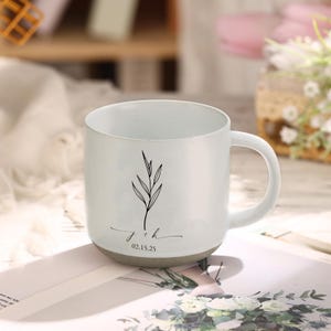 May include: White ceramic mug with a black botanical design and the initials "j + h" with the date "02.15.25" in a script font. The mug has a speckled finish and a gray base, perfect for coffee or tea.