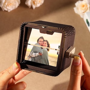 May include: A dark brown wooden photo cube with a picture of two people embracing. The cube has a crank on the side. The photo cube is held by a person's hands. A unique gift idea.
