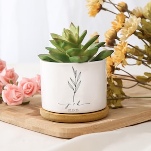 Op de afbeelding: Een witte keramische plantenbak met een groene vetplant. De plantenbak heeft een zwart bloemmotief en de initialen "j + h" met de datum "02.15.25". De plantenbak staat op een ronde, natuurlijke houten onderzetter.