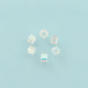 May include: Six clear, iridescent cube-shaped beads arranged in a circle on a light blue background. The beads have a subtle rainbow sheen, reflecting various colors. The beads are small and appear to be made of glass or crystal.
