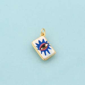 May include: Gold-toned rectangular pendant with a white background, featuring a blue and red eye design. The pendant has a gold loop for attaching to a chain or bracelet. The eye design is centered on the pendant.