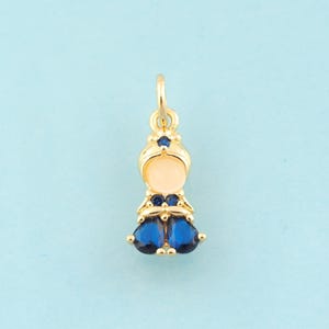 May include: Gold charm with a blue and white enamel doll design. The doll has a blue crown and blue dress with blue gem accents.