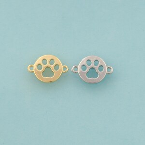 Puede incluir: Dos conectores de abalorios metálicos, uno dorado y otro plateado, cada uno con un diseño de huella de pata dentro de un marco circular. Los abalorios tienen pequeños bucles para sujetarlos a la joyería. El fondo es azul claro.