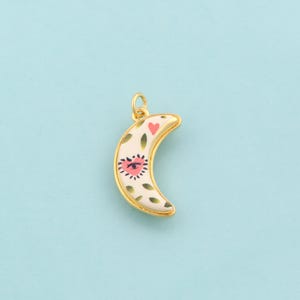 May include: Gold-toned crescent moon pendant with a cream-colored enamel finish. The pendant features a pink heart and eye design, along with green leaf accents. The pendant is approximately 1 inch in length.
