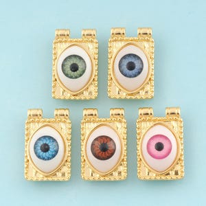 May include: Five gold-toned eye charms with different colored irises. Each charm is a rectangle with a white background and a black outline. The irises are blue, green, brown, pink, and a mix of blue and green.