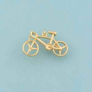 May include: A gold-colored bicycle charm with a detailed design. The charm features a classic bicycle silhouette with wheels, handlebars, a seat, and a chain. The charm is suspended from a small loop, ready to be added to a necklace or bracelet.