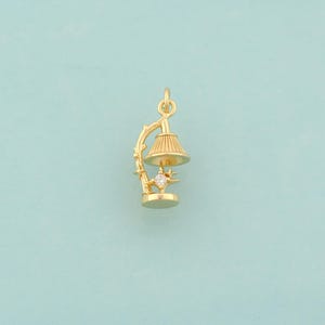 May include: A gold-colored charm in the shape of a small lamp. The lamp has a detailed lampshade, a decorative stem, and a starburst design with a small, clear gemstone. The charm is designed to be attached to a necklace or bracelet.