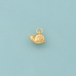 Peut inclure: Un charm en forme d'escargot doré, avec une coquille détaillée et une boucle pour l'attacher à un collier ou un bracelet. L'escargot a un visage souriant et est posé sur un fond bleu clair. Ce charm est idéal pour la création de bijoux.