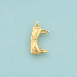 May include: A gold-colored pendant in the shape of a bra. The pendant has a curved rectangular body with a textured surface and two pointed projections. The pendant is suspended from a small loop.