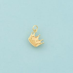 Op de afbeelding: Een goudkleurige kroonbedel met een lus om aan een ketting of armband te bevestigen. De kroon heeft drie punten en is versierd met kleine, ronde details. De achtergrond is effen lichtblauw.