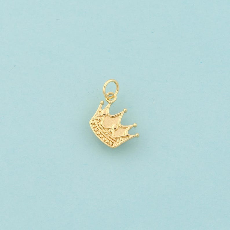 Op de afbeelding: Een goudkleurige kroonbedel met een lus om aan een ketting of armband te bevestigen. De kroon heeft drie punten en is versierd met kleine, ronde details. De achtergrond is effen lichtblauw.