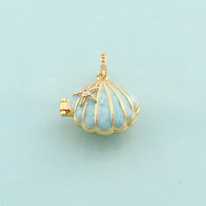 May include: A gold-toned seashell pendant with a light blue enamel finish. The pendant features a small starfish with a clear crystal in the center. The seashell opens to reveal a small compartment. The pendant hangs from a gold-toned bail.