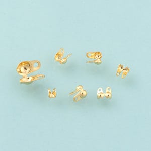 May include: A collection of small, gold-colored metal clasps, each with a unique design. Some have a loop and a ball, while others have a more intricate shape. The clasps are scattered against a light blue background.