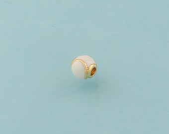 Puede incluir: Una pequeña cuenta redonda con un acabado de esmalte blanco y detalles dorados. La cuenta tiene un diseño de pelota de tenis con una banda dorada alrededor del centro. La cuenta tiene un agujero para ensartar. El fondo es azul claro liso.
