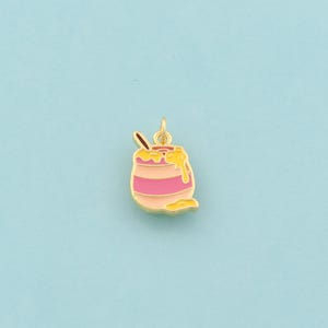 Puede incluir: Un dije dorado con forma de tarro de miel con esmalte rosa y amarillo. El tarro de miel tiene una cuchara y miel goteando. El dije está sobre un fondo azul claro.