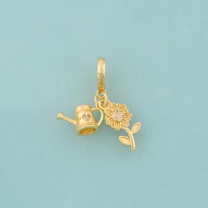 Puede incluir: Charm dorado con una regadera y un girasol. La regadera tiene las palabras "water for bloom" y un pequeño diseño floral. El girasol tiene un centro detallado y está unido a un tallo con hojas. El charm tiene un cierre.