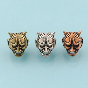 Op de afbeelding: Drie metalen bedels in de vorm van een Japans Oni-masker. De maskers zijn goud, zilver en koper.