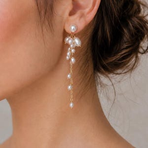 May include: A gold-toned chain earring featuring a cluster of white freshwater pearls at the earlobe, with a delicate dangling chain extension adorned with several smaller, evenly spaced white pearls hanging vertically from the main cluster.