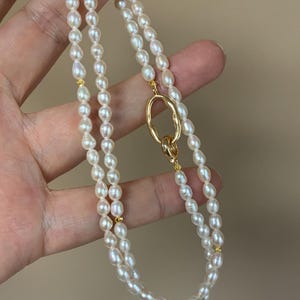 Puede incluir: Un collar de perlas de doble hebra con un cierre ovalado dorado. El collar presenta pequeñas perlas blancas de forma ovalada. Se ven detalles dorados entre las hebras de perlas. El collar se muestra sobre un fondo neutro.