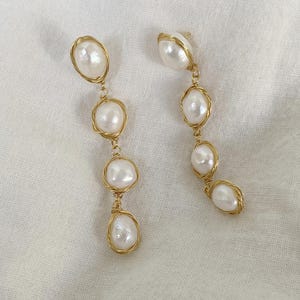 Puede incluir: Un par de pendientes de alambre dorado con cuatro perlas blancas cada uno. Las perlas están enhebradas en el alambre y son de diferentes tamaños.