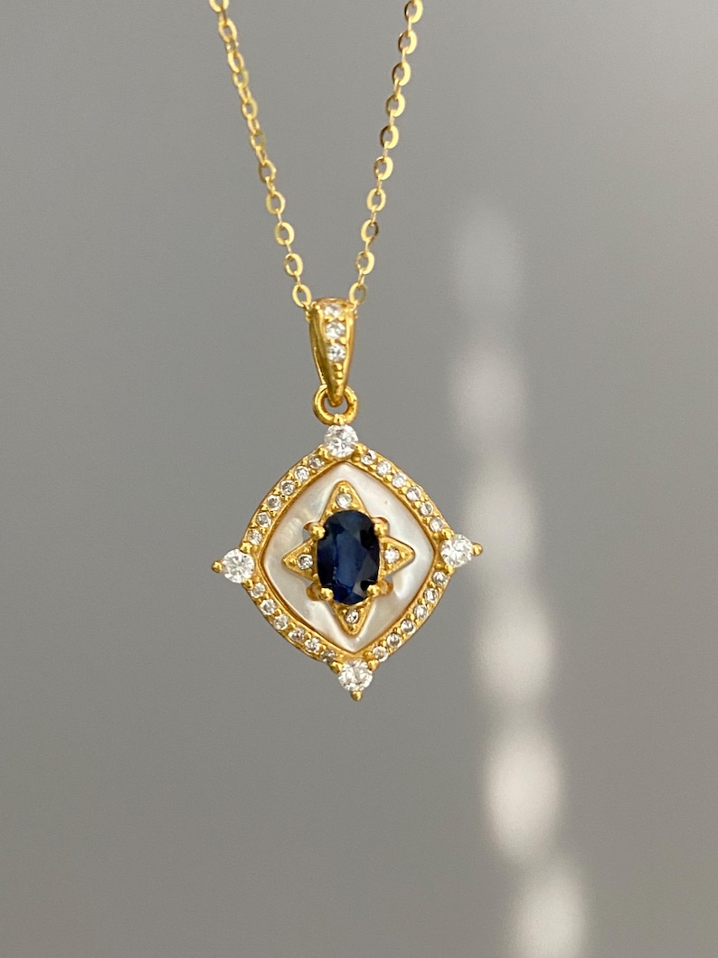 May include: A gold-colored pendant necklace featuring a central oval-shaped dark blue gemstone. The pendant is set within a white square-shaped frame, surrounded by small clear gemstones and a gold-colored border. The necklace chain is also gold-colored.