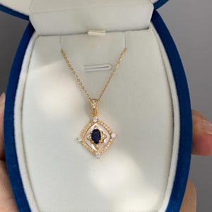 May include: A gold necklace with a pendant. The pendant has a dark blue gemstone in a white and gold setting, with small, clear stones around the edges. The necklace is displayed in a blue jewelry box.