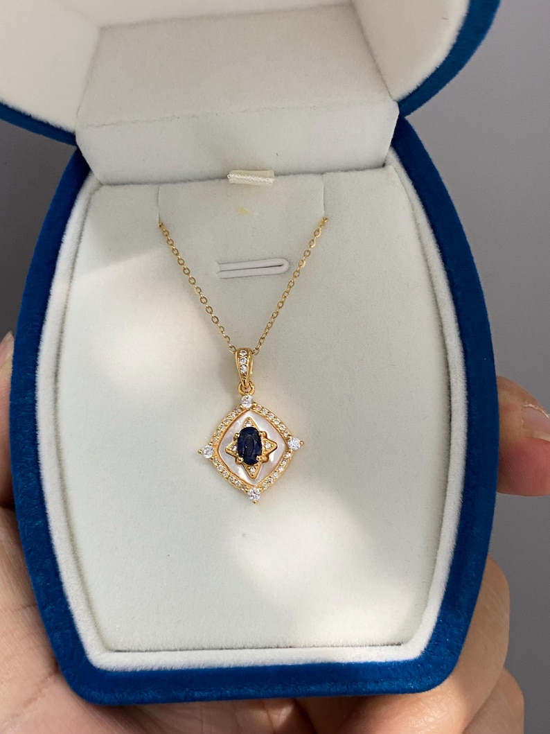 May include: A gold necklace with a pendant. The pendant has a dark blue gemstone in a white and gold setting, with small, clear stones around the edges. The necklace is displayed in a blue jewelry box.