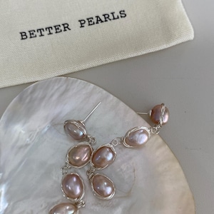 May include: A pair of dangle earrings featuring oval-shaped, light pink pearls wrapped in silver wire. The earrings are displayed on a white, shell-shaped surface, with a small cloth bag that reads "BETTER PEARLS" in black text.