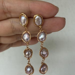 Puede incluir: Un par de pendientes colgantes con tres perlas barrocas de color púrpura claro cada uno, encerradas en alambre dorado. Los pendientes cuelgan de un lazo de alambre dorado. Las perlas tienen un brillo lustroso.