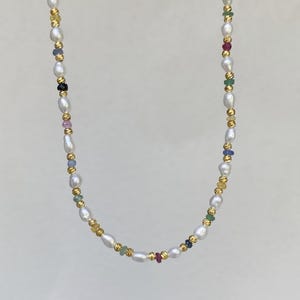 Puede incluir: Un delicado collar con perlas de agua dulce blancas intercaladas con pequeñas piedras preciosas de colores y cuentas doradas. El collar tiene un diseño sencillo, con las perlas y las piedras preciosas ensartadas juntas.