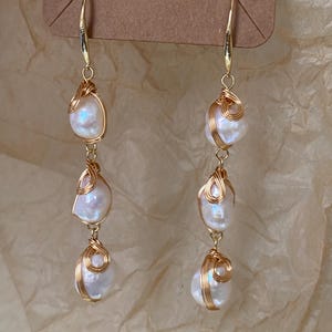 Puede incluir: Pendientes de alambre dorado con tres perlas iridiscentes en forma de lágrima. Las perlas cuelgan de un gancho dorado, creando un efecto colgante. Los pendientes se muestran sobre un fondo neutro y texturizado.