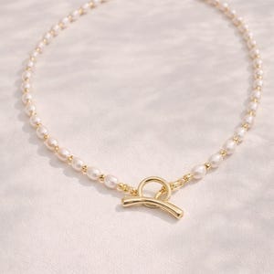 Pode incluir: Um delicado colar de pérolas com fecho dourado. O colar apresenta pequenas pérolas brancas enfiadas com detalhes dourados. O fecho é um design de barra e anel dourado.