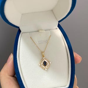 May include: A gold necklace with a pendant featuring a dark blue gemstone at the centre, surrounded by small white stones and a white border. The necklace is presented in a blue velvet jewellery box.