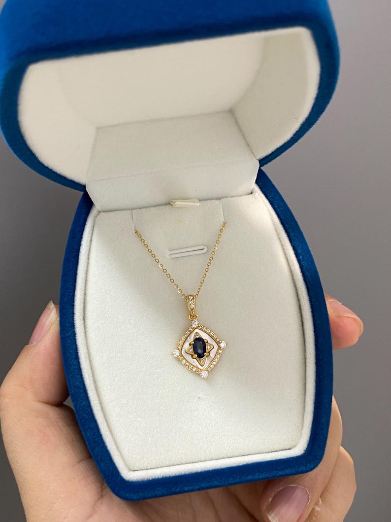 May include: A gold necklace with a pendant featuring a dark blue gemstone at the centre, surrounded by small white stones and a white border. The necklace is presented in a blue velvet jewellery box.