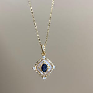 May include: A gold necklace with a pendant. The pendant has a dark blue oval gemstone at its centre, surrounded by a white border and small clear gemstones, set in a square shape. The chain is delicate and gold-coloured.