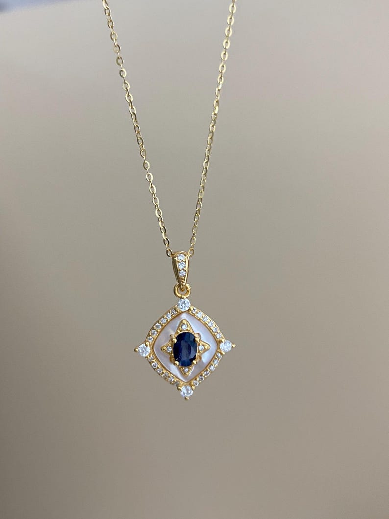May include: A gold necklace with a pendant. The pendant has a dark blue oval gemstone at its centre, surrounded by a white border and small clear gemstones, set in a square shape. The chain is delicate and gold-coloured.