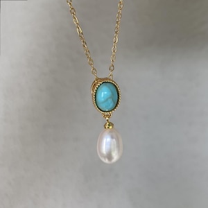 May include: A gold-colored chain necklace with a pendant featuring a turquoise-colored oval stone set in a gold frame. Below the stone hangs a white, teardrop-shaped pearl. The necklace is displayed against a soft, neutral background.